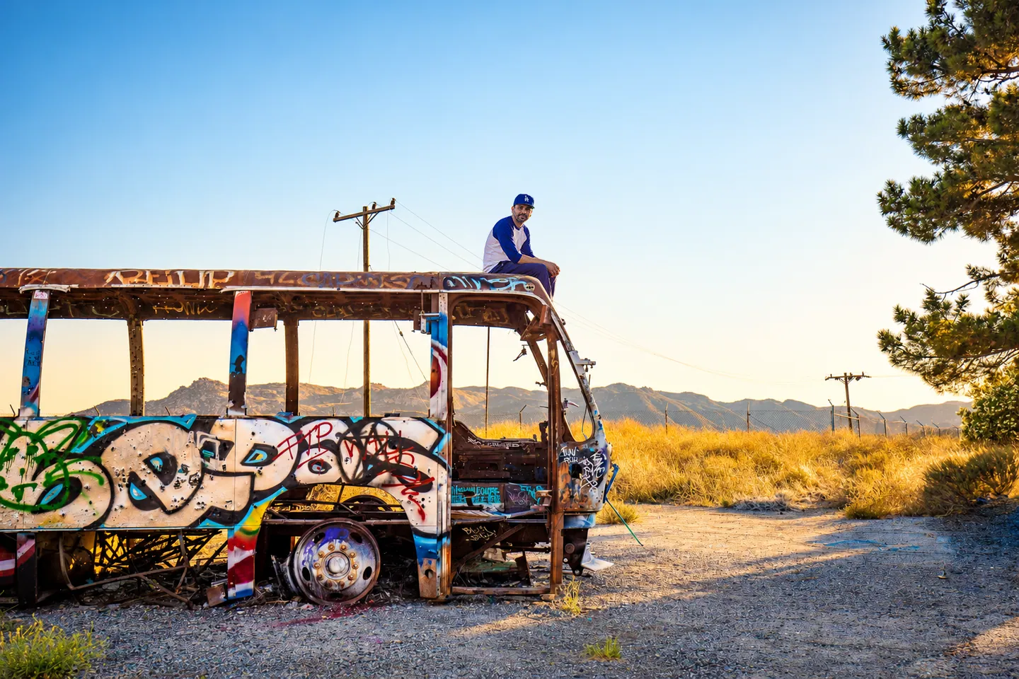 Marco sitting on the roof of a graffiti-covered bus in the desert, wearing a Dodgers cap.